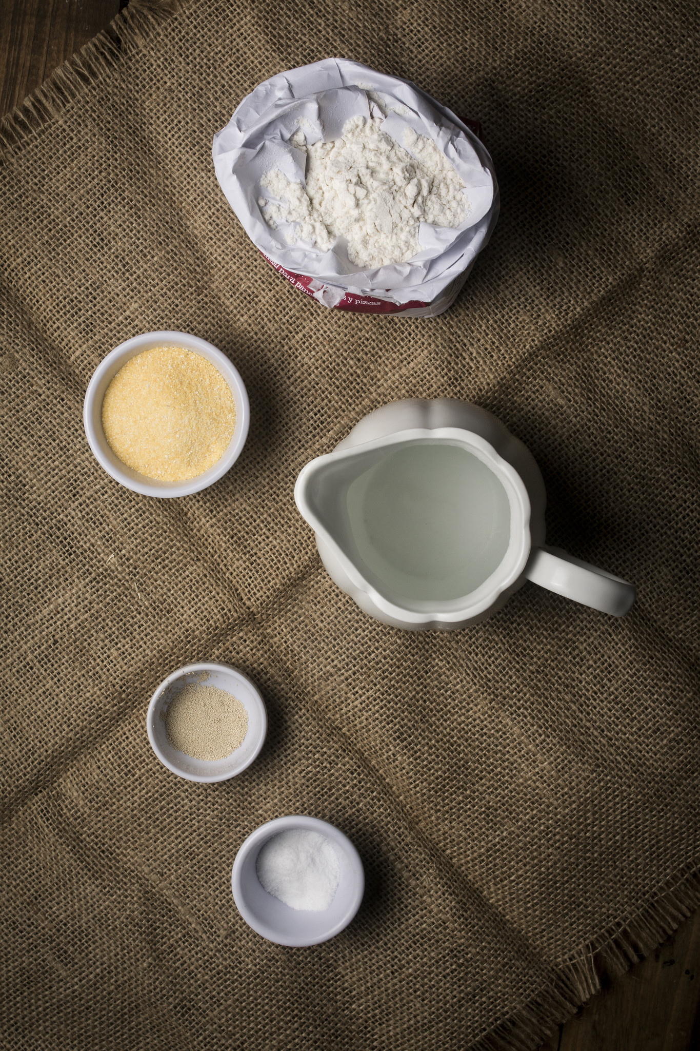 Ingredients for no-knead ciabatta rolls including bread flour, water, yeast, salt and semolina on a rustic cloth.